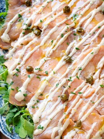 Salmon carpaccio on a serving plate garnished with capers, parsley and lemon zest.